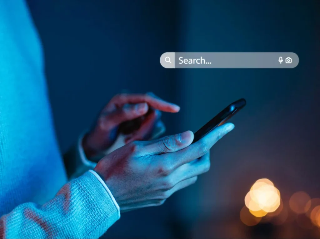 A person using a smartphone with a search bar displayed on the screen in a dark setting.