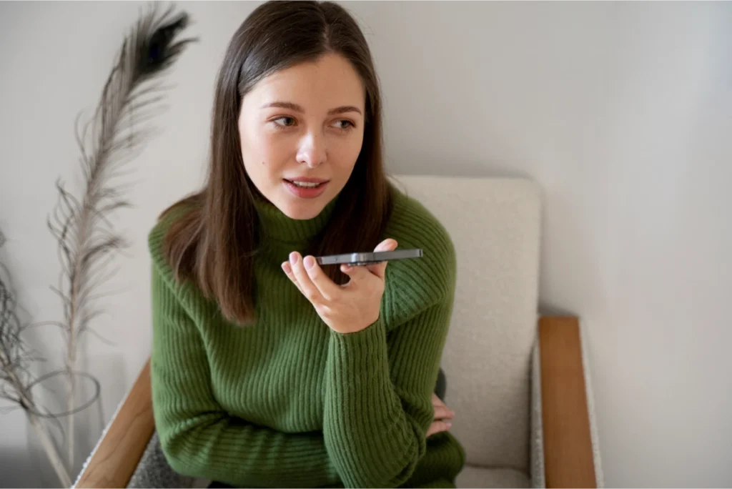 Woman in a green sweater speaking into a smartphone on speaker mode while sitting on a beige armchair indoors