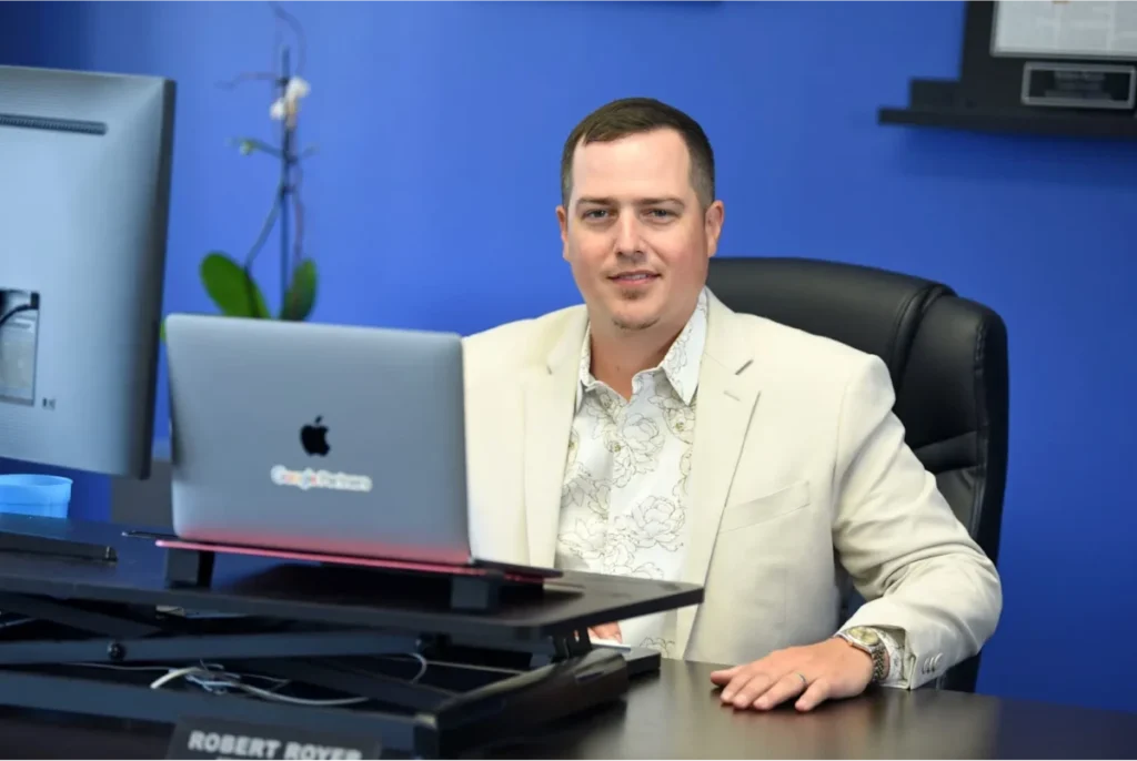 Man in a beige blazer sitting at a desk with a laptop and desktop monitor, smiling in an office with a blue wall background.