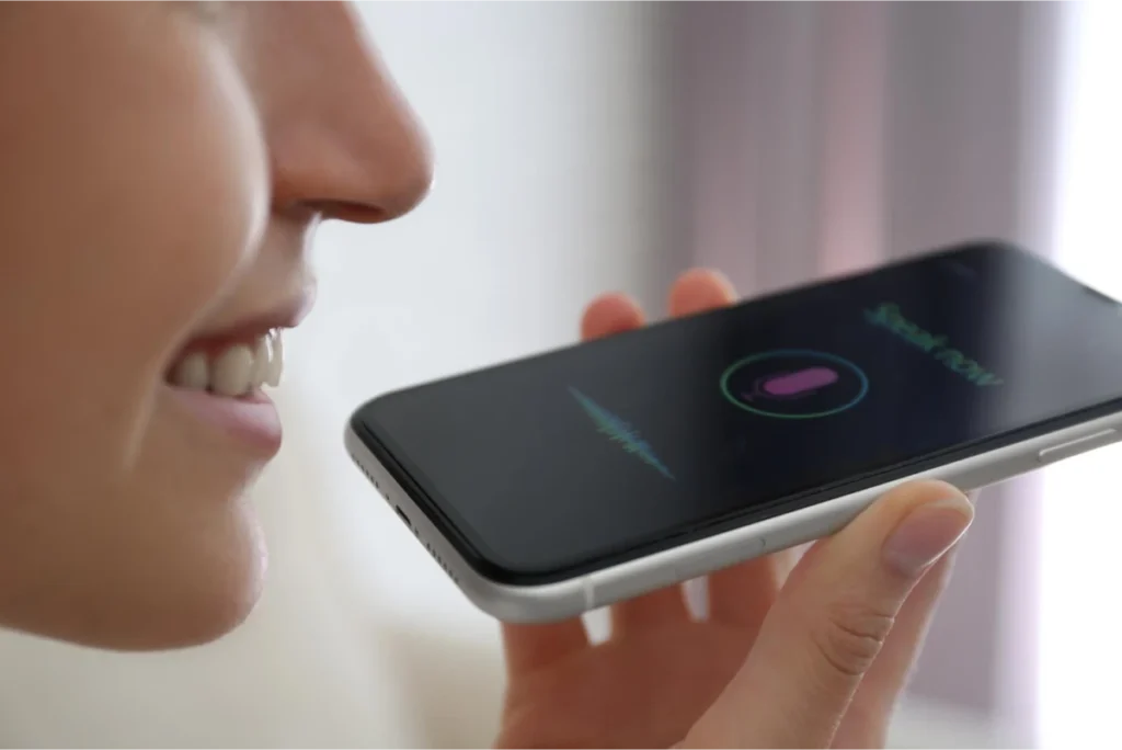 Close-up of a woman using voice command on a smartphone, speaking into the device with a voice assistant interface displayed on the screen.