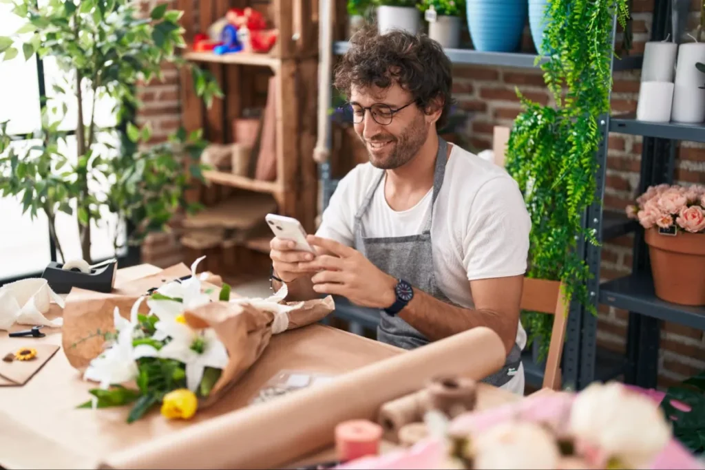 A smiling florist wearing glasses and a gray apron uses a smartphone while sitting at a wooden table with flower arrangements, wrapping paper, and tools in a cozy flower shop.