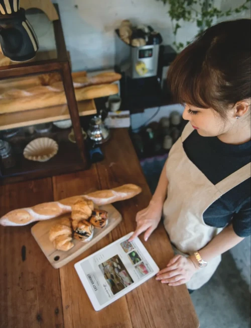 A woman wearing a beige apron stands at a wooden counter with croissants and a baguette on a cutting board. She is using a tablet, which displays images of baked goods, inside a cozy bakery setting.